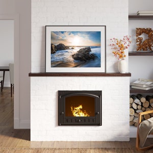 May include: A white brick fireplace with a black cast iron wood-burning stove. The stove has a glass door and a fire burning inside. Above the stove is a framed picture of a beach scene with a sunset. There is a wooden shelf above the picture and a rocking chair with a blanket on it to the right.
