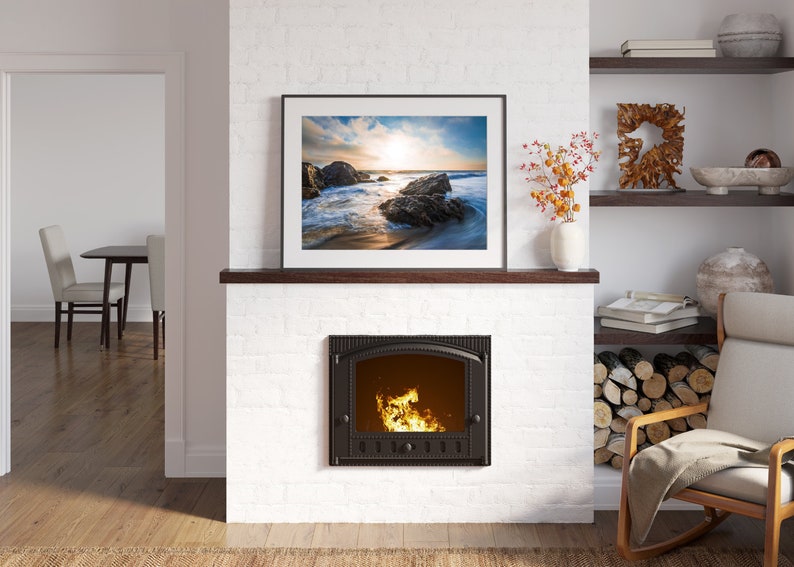May include: A white brick fireplace with a black cast iron wood-burning stove. The stove has a glass door and a fire burning inside. Above the stove is a framed picture of a beach scene with a sunset. There is a wooden shelf above the picture and a rocking chair with a blanket on it to the right.