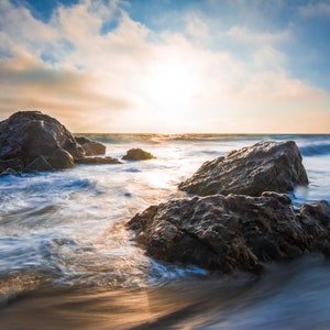 May include: A scenic view of a rocky beach with waves crashing against the shore. The sun is setting in the distance, casting a golden glow over the water. The rocks are dark and rugged, and the water is a mix of blue and white.