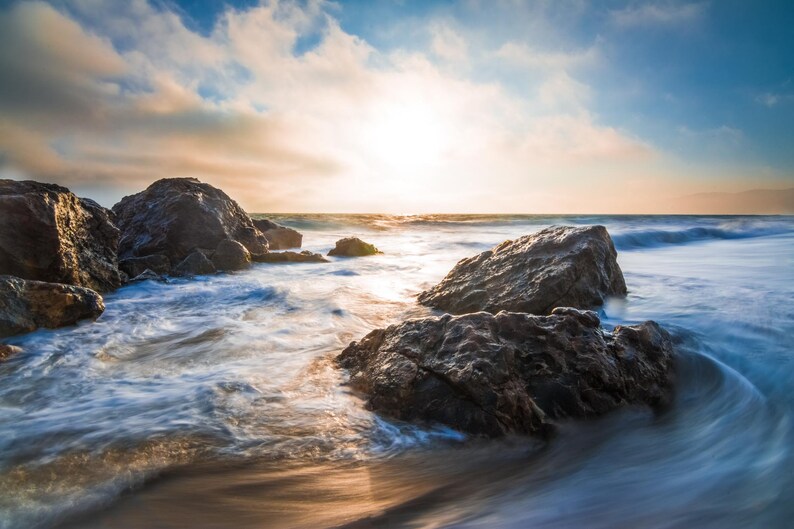 May include: A scenic view of a rocky beach with waves crashing against the shore. The sun is setting in the distance, casting a golden glow over the water. The rocks are dark and rugged, and the water is a mix of blue and white.