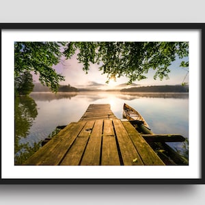 Op de afbeelding: Een houten steiger steekt uit over een kalm meer met een kleine houten boot vastgemaakt aan het einde. Het meer is omgeven door bomen en de zon schijnt door de takken.