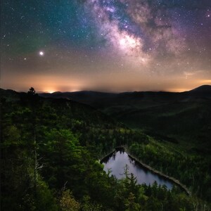 May include: A night sky with the Milky Way galaxy visible above a dark forest with a small lake in the center.