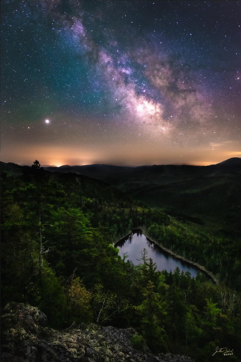 May include: A night sky with the Milky Way galaxy visible above a dark forest with a small lake in the center.