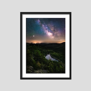 May include: A night sky with the Milky Way galaxy visible above a dark green forest and a small lake. The stars are bright and the sky is a deep blue.