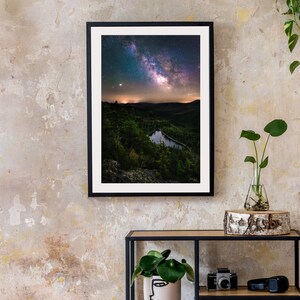 May include: A framed photograph of a night sky with the Milky Way galaxy visible above a dark green forest and a small lake. The photo is hanging on a light beige wall with a wooden shelf and a plant in a glass vase below it.