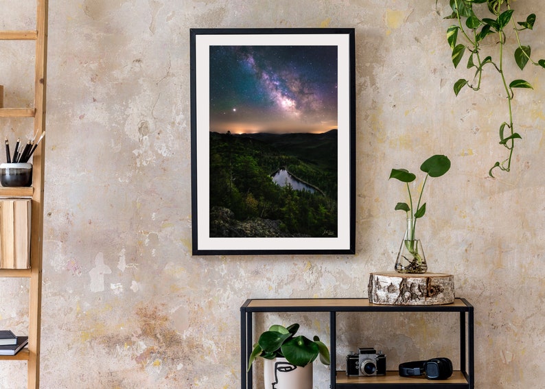 May include: A framed photograph of a night sky with the Milky Way galaxy visible above a dark green forest and a small lake. The photo is hanging on a light beige wall with a wooden shelf and a plant in a glass vase below it.