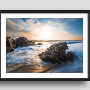 May include: A scenic photograph of a rocky beach with waves crashing against the shore. The sun is setting in the distance, casting a golden glow over the water. The sky is a mix of blue and white clouds.