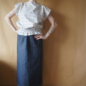 Pode incluir: Uma mulher vestindo uma blusa branca e rosa xadrez com uma bainha franjada e uma saia jeans azul escura. Ela tem cabelo ruivo e est&aacute; em p&eacute; em frente a uma parede marrom.