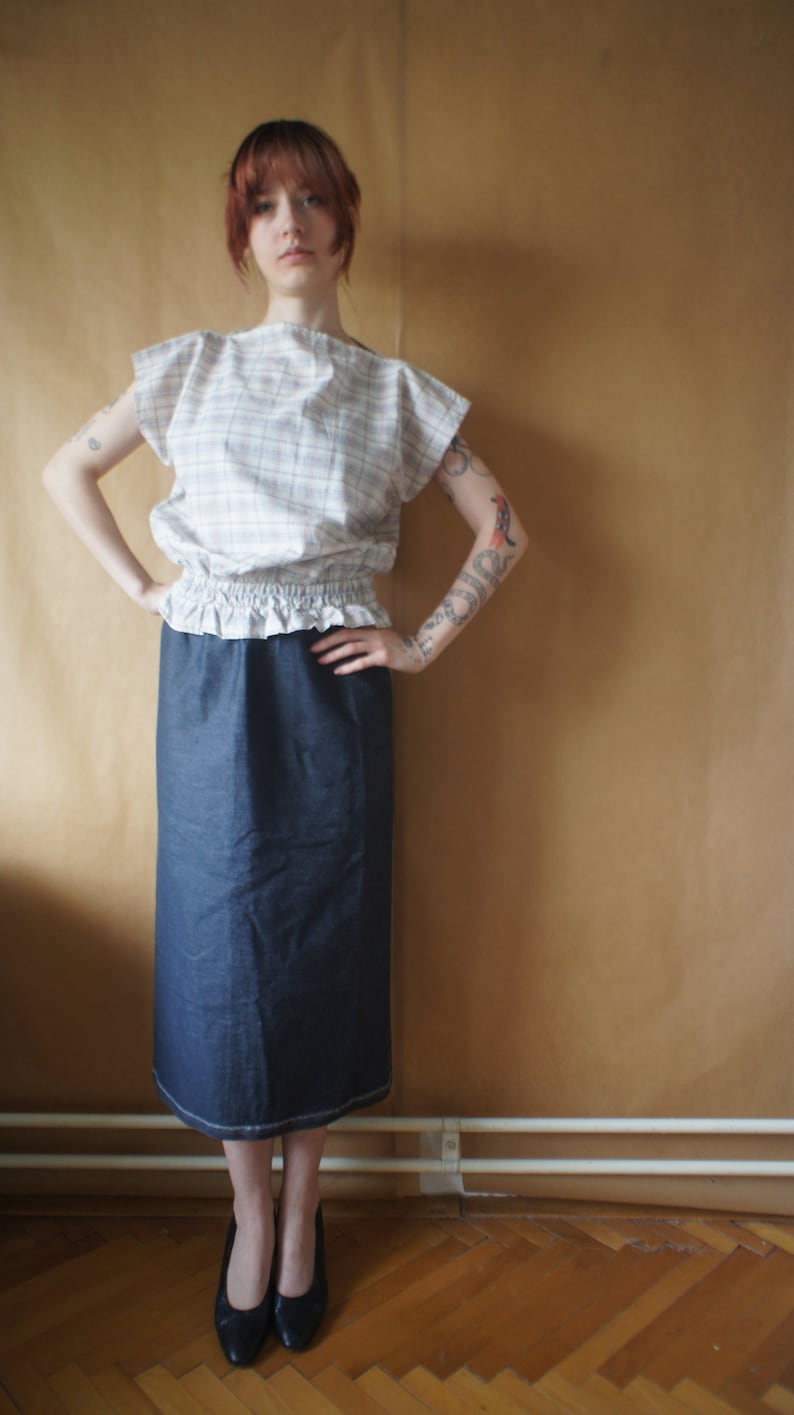 Pode incluir: Uma mulher vestindo uma blusa branca e rosa xadrez com uma bainha franjada e uma saia jeans azul escura. Ela tem cabelo ruivo e est&aacute; em p&eacute; em frente a uma parede marrom.