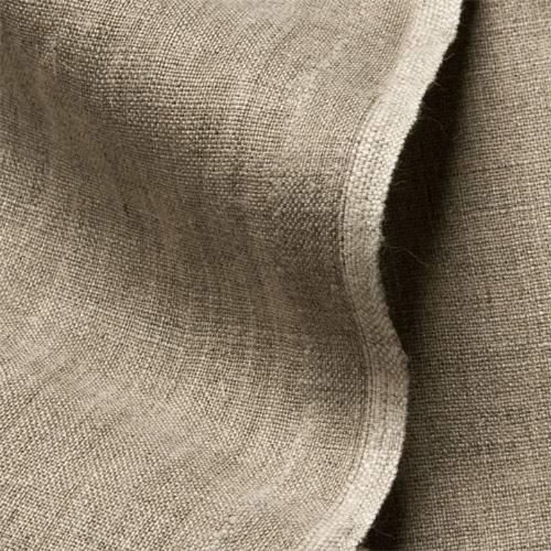 May include: Close-up of a beige linen fabric with a textured weave. The fabric is folded, creating shadows and highlighting the natural fibers. The neutral color and texture suggest a focus on natural textiles.