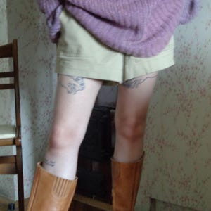 Pu&ograve; includere: Una persona indossa un maglione lilla, pantaloncini color cachi e stivali di pelle marrone. Gli stivali arrivano al ginocchio e hanno una cerniera. I pantaloncini sono a vita alta. Lo sfondo ha una carta da parati floreale.