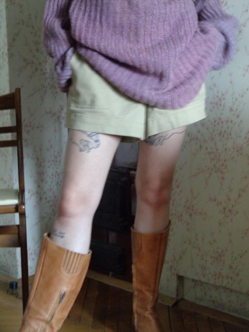 Pu&ograve; includere: Una persona indossa un maglione lilla, pantaloncini color cachi e stivali di pelle marrone. Gli stivali arrivano al ginocchio e hanno una cerniera. I pantaloncini sono a vita alta. Lo sfondo ha una carta da parati floreale.