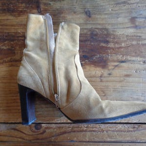 May include: A tan suede ankle boot with a pointed toe and a dark brown block heel. The boot features a side zipper and a slightly curved topline. The boot is set against a wooden background.