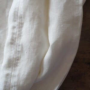 May include: Close-up of off-white linen fabric with a visible seam. The fabric is folded, creating soft curves and shadows. The texture of the linen is apparent, suggesting a natural, breathable material. The overall aesthetic is simple and elegant.