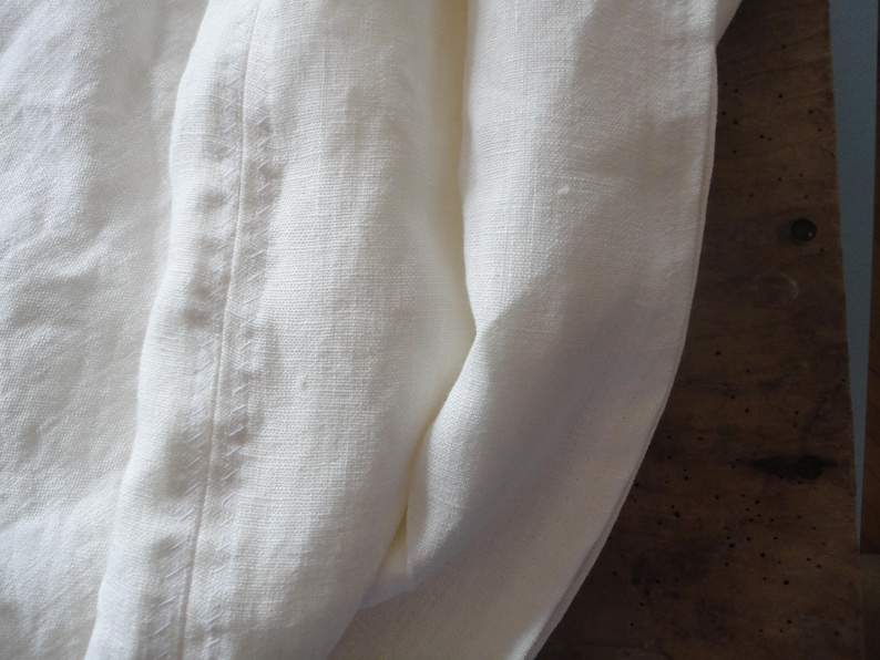May include: Close-up of off-white linen fabric with a visible seam. The fabric is folded, creating soft curves and shadows. The texture of the linen is apparent, suggesting a natural, breathable material. The overall aesthetic is simple and elegant.