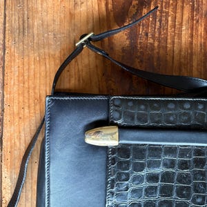 Può includere: Borsa a tracolla in pelle nera con chiusura e attacchi per cinturino color oro. La borsa presenta un motivo strutturato, simile alla pelle di coccodrillo, sul pannello frontale e finiture in pelle liscia sui lati. Il cinturino è sottile e regolabile.