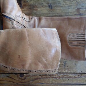 Pu&ograve; includere: Un paio di stivali da equitazione in pelle marrone chiaro. Gli stivali hanno una cerniera, cuciture decorative e una punta arrotondata. La pelle ha un aspetto usurato. Gli stivali sono su una superficie di legno.