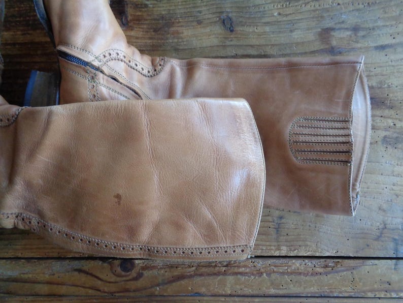 Pu&ograve; includere: Un paio di stivali da equitazione in pelle marrone chiaro. Gli stivali hanno una cerniera, cuciture decorative e una punta arrotondata. La pelle ha un aspetto usurato. Gli stivali sono su una superficie di legno.