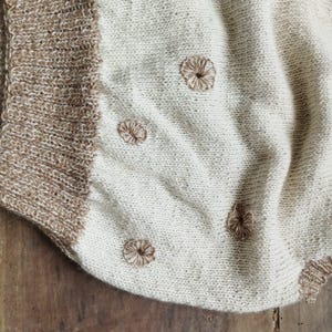 以下が含まれることがあります： クリーム色のニットブランケット、茶色のトリムと刺繍された花のアクセント。 ブランケットは、テクスチャードステッチパターンと柔らかく魅力的な外観が特徴です。 茶色のトリムは端を縁取り、花のアクセントは均等に配置されています。