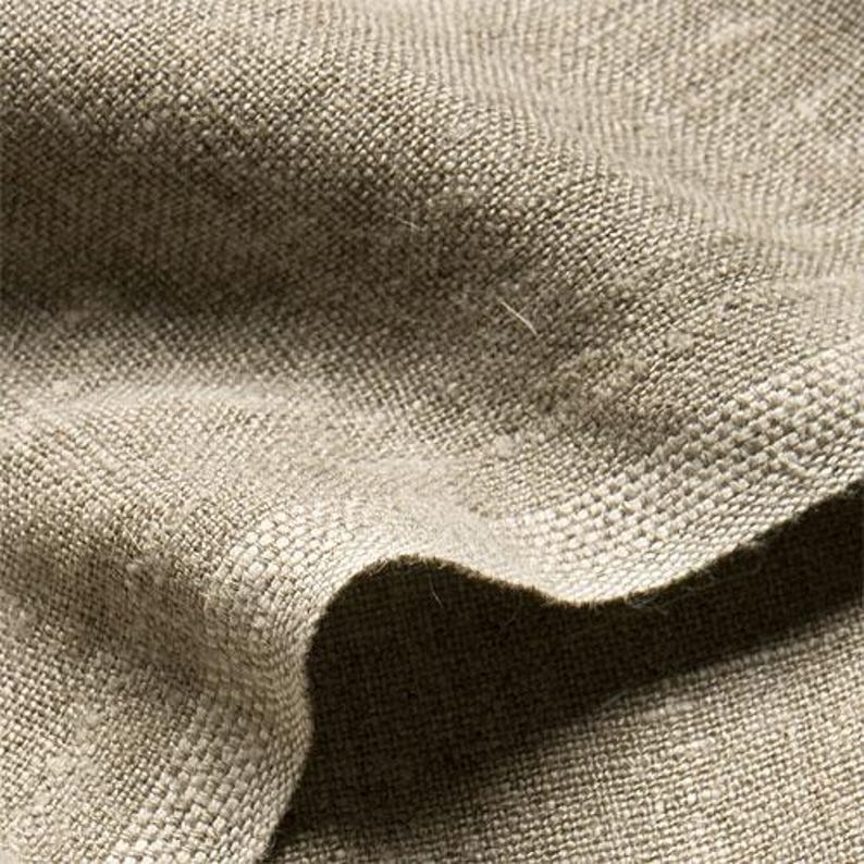 May include: Close-up of a textured, beige linen fabric. The weave is visible, with a slightly irregular pattern. The fabric is folded, creating shadows and highlighting the texture. The color is a neutral, earthy tone.