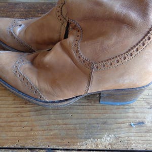 Pu&ograve; includere: Un paio di stivali in pelle marrone chiaro con perforazioni decorative e un tacco basso. Gli stivali hanno un aspetto usurato e vintage, con pieghe e segni di usura visibili. Gli stivali sono su una superficie di legno.