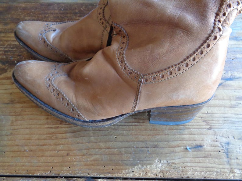 Pu&ograve; includere: Un paio di stivali in pelle marrone chiaro con perforazioni decorative e un tacco basso. Gli stivali hanno un aspetto usurato e vintage, con pieghe e segni di usura visibili. Gli stivali sono su una superficie di legno.