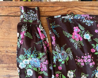 May include: Close-up of a brown fabric with a floral pattern. The design features pink, blue, and white flowers with green foliage. The fabric is likely part of a garment, with a gathered sleeve detail visible.