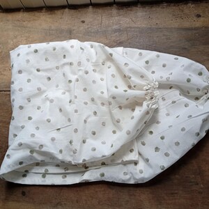 May include: A white fabric garment featuring a repeating pattern of muted sage green polka dots. The item has gathered, elasticized openings at the ends, creating a soft, draped silhouette with visible folds throughout the textile material.