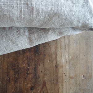 May include: Close-up of folded, light gray linen fabric on a weathered, brown wooden surface. The fabric has a textured appearance, with visible weave details. The wood grain and knots add a rustic aesthetic.