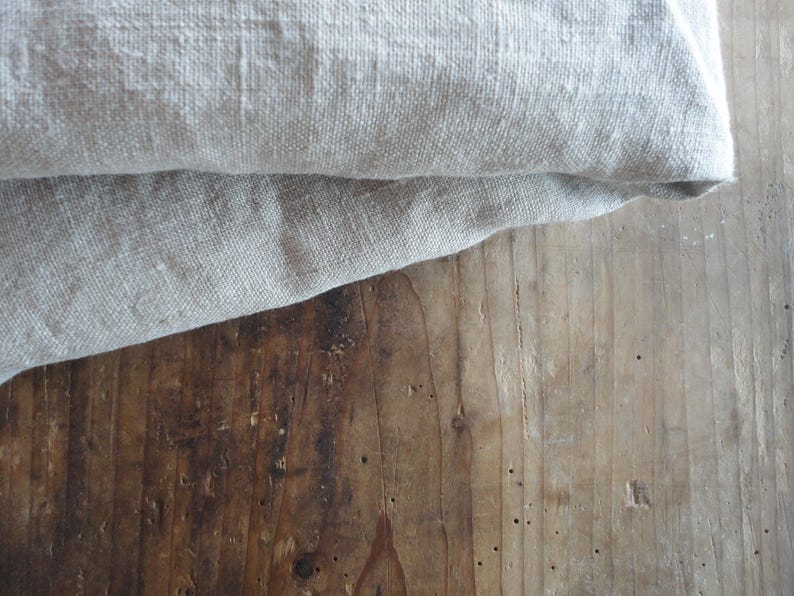 May include: Close-up of folded, light gray linen fabric on a weathered, brown wooden surface. The fabric has a textured appearance, with visible weave details. The wood grain and knots add a rustic aesthetic.