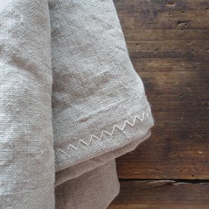 May include: Close-up of a folded, light gray linen fabric with a white zigzag stitched edge. The fabric has a textured appearance and is set against a dark brown wooden surface.