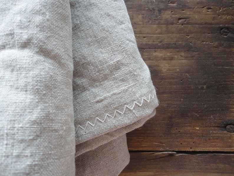 May include: Close-up of a folded, light gray linen fabric with a white zigzag stitched edge. The fabric has a textured appearance and is set against a dark brown wooden surface.