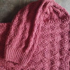 May include: A dusty rose knitted sweater with a hood and long sleeves. The sweater has a textured knit pattern and ribbed cuffs. The garment is made of a soft material, possibly wool.