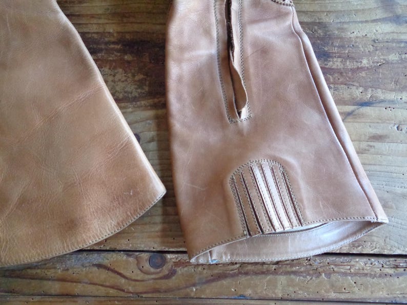 Pu&ograve; includere: Primo piano di due chaps da equitazione in pelle marrone chiaro. Gli chaps hanno una superficie strutturata e cuciture decorative. Uno chap ha un pannello decorativo con linee verticali nella parte inferiore. Sfondo in legno.