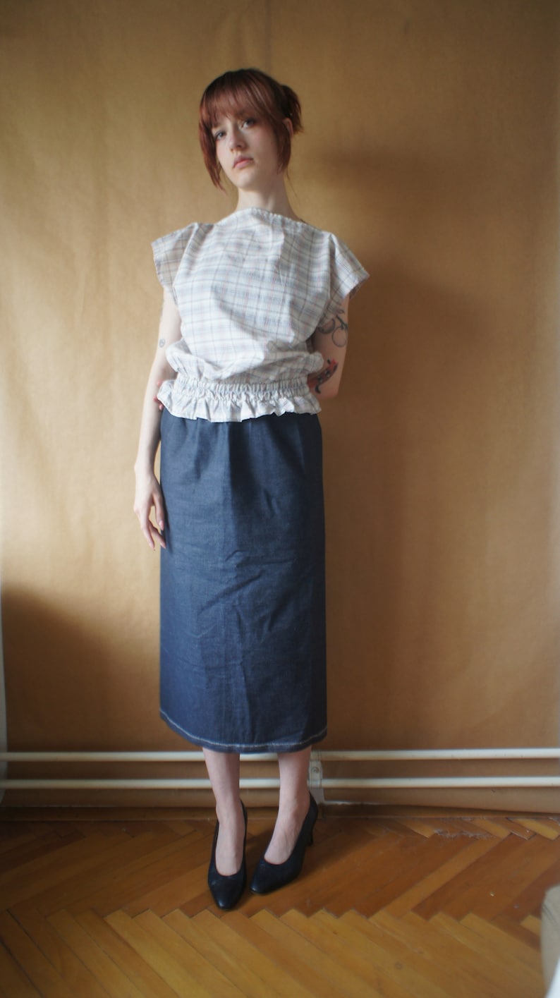 Pode incluir: Uma mulher vestindo uma blusa branca e rosa xadrez com uma bainha franjada e uma saia jeans azul escura. Ela est&aacute; usando sapatos pretos de salto alto.