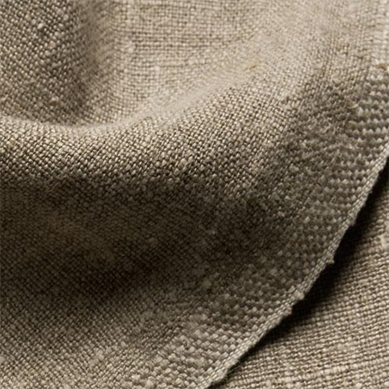 May include: Close-up of a textured, woven fabric in shades of tan and beige. The fabric has a coarse, irregular weave, with visible fibers and a slightly uneven surface. The folds and shadows create depth and texture.