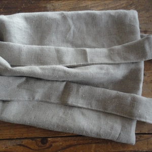 Może przedstawiać: Złożona, jasno szara tkanina lniana z dwoma długimi, przymocowanymi paskami. Tkanina ma lekko teksturowany wygląd i leży na drewnianej powierzchni. Paski są złożone na głównym korpusie tkaniny.