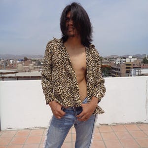 Puede incluir: Una camisa con estampado de leopardo y vaqueros azules con flecos. La camisa está desabrochada. Los vaqueros son de mezclilla azul claro. El fondo muestra un paisaje urbano bajo un cielo nublado.