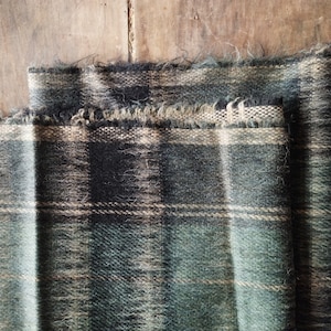 Puede incluir: Primer plano de una tela a cuadros doblada en tonos verdes, negros y beige. La tela tiene un aspecto texturizado con hilos tejidos visibles y un borde deshilachado. El patrón de cuadros presenta una cuadrícula de líneas que se cruzan.