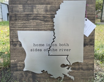 May include: A wooden sign with a metal cutout of the state of Louisiana. The metal is silver and the wood is dark brown. The text "home is on both sides of the river" is printed on the metal cutout.