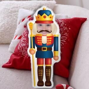 May include: A white pillow shaped like a nutcracker with a child's face in the middle. The nutcracker is wearing a blue and red uniform with a gold crown and holding a gold staff.
