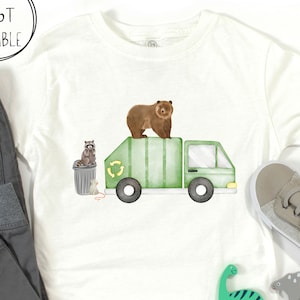 May include: A white short-sleeve t-shirt featuring a graphic of a green garbage truck with a brown bear on top, a raccoon on a trash can, and a small mouse. The text SIZES 6M-6T AVAILABLE is printed in a circle in the upper left corner.