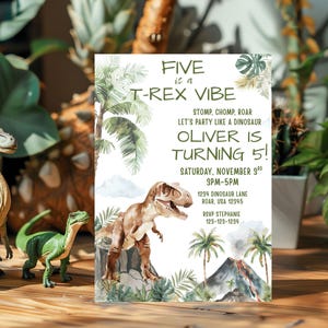 Pode incluir: Um convite de anivers&aacute;rio com tema de dinossauro com o texto "FIVE is a T-REX VIBE" e "OLIVER IS TURNING 5!". O convite apresenta uma ilustra&ccedil;&atilde;o em aquarela de um T-Rex, palmeiras e um vulc&atilde;o. Estatuetas de dinossauros em primeiro plano.