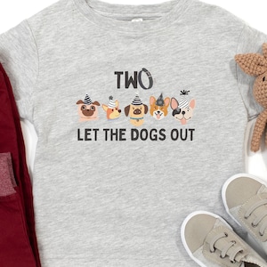 May include: Heather gray toddler t-shirt with the text "TWO LET THE DOGS OUT" and a graphic of six dogs wearing party hats. The shirt is paired with red pants, gray sneakers, and a brown stuffed animal.