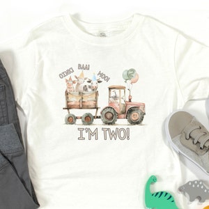 May include: White short-sleeved t-shirt with a watercolor illustration of a red tractor pulling a wagon with farm animals. The animals are a pig, a cow, and a calf. The text "OINK! BAA! MOO! I'M TWO!" is printed on the shirt.