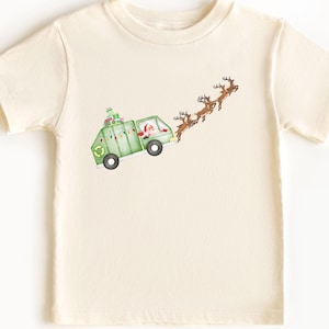 May include: A cream-colored short-sleeve t-shirt featuring a graphic illustration of a green garbage truck driven by Santa Claus, carrying wrapped gifts on the roof and being pulled through the air by a team of brown reindeer in flight.