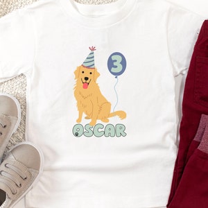 May include: White toddler t-shirt featuring a golden retriever wearing a party hat and holding a blue balloon with the number 3. The name "OSCAR" is printed below the dog in teal letters. The word "White" is printed at the bottom of the shirt.