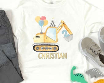 May include: A white t-shirt featuring a yellow construction excavator graphic wearing a striped party hat, carrying a blue number two in its bucket, and holding colorful balloons. The name CHRISTIAN is printed below in yellow capital letters.