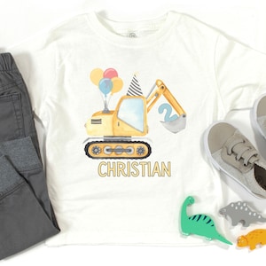 May include: A white t-shirt with a yellow excavator design, balloons, and the number 2. The text "CHRISTIAN" is printed below the excavator.
