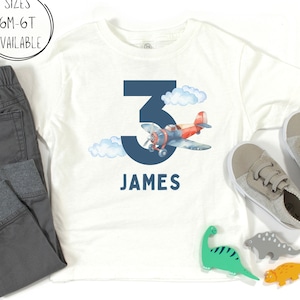 May include: White t-shirt with a navy blue number 3, an airplane graphic, and the name "JAMES" in navy blue. The shirt is paired with gray pants and sneakers. Sizes 6M-6T available.